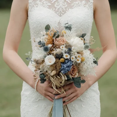 Boho style flowers bridal bouquet with ivory roses, dusty blue blooms, eucalyptus, white dried ferns, golden accents, and beige preserved flowers.