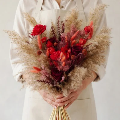 Boho style flowers bridal bouquet with burgundy roses, crimson blooms, coral dried grasses, dusty foliage, and large beige pampas plumes.