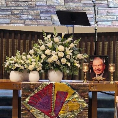 Celebration of life flowers set with white roses, lilies, snapdragons, and greenery arranged beside a memorial photo and candles.